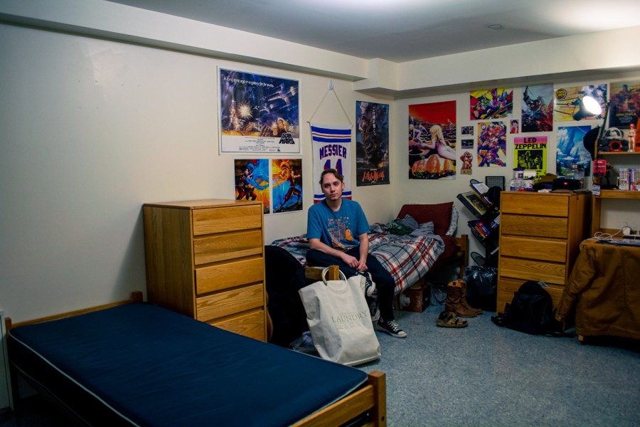 A man sits on his bed looking at the camera in front of his decorated walls next to an empty bed and drawer that is not being used.