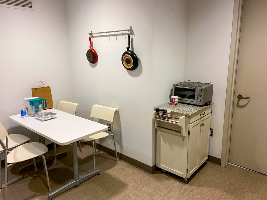 A multipurpose dining and cooking space in a residence hall on NYU’s Washington, D.C., campus. On the left, there’s a white table with four white chairs tucked underneath. On the right, next to the door, is a mobile island with a microwave.
