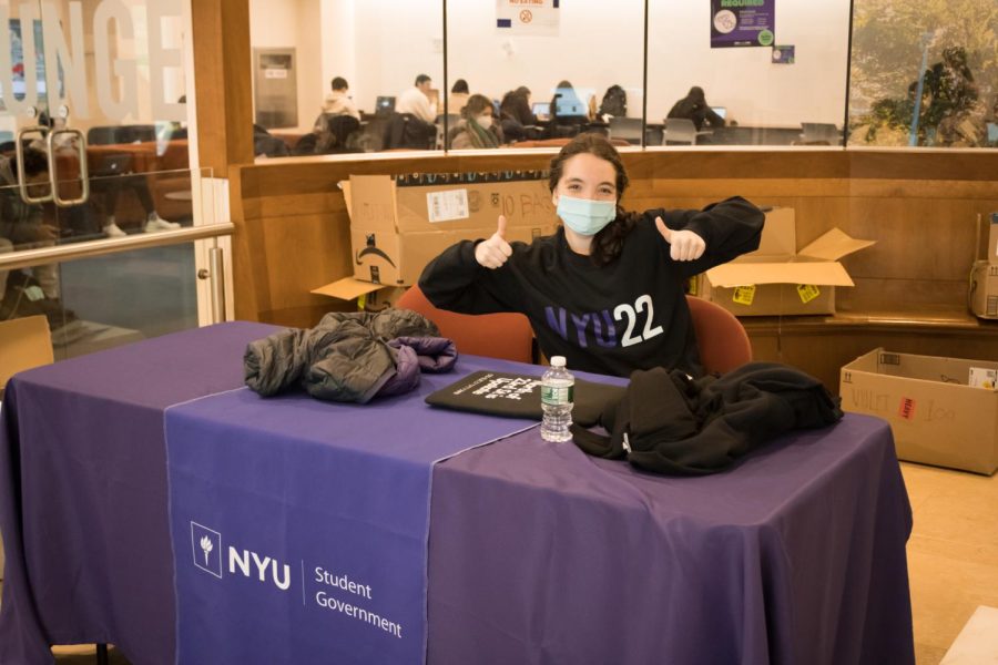 At the Kimmel Center for University Life, a girl sits at a table with a purple tablecloth and a sign that reads “NYU Student Government”. She holds two thumbs up, and behind her there are cardboard boxes and the Commuter Lounge is visible in the background.