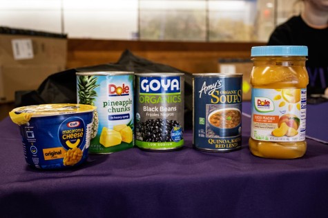 On top of a table with a purple tablecloth, there is from left to right: Kraft macaroni and cheese, canned pineapple chunks, canned black beans, canned lentils and sliced peaches in a plastic container.