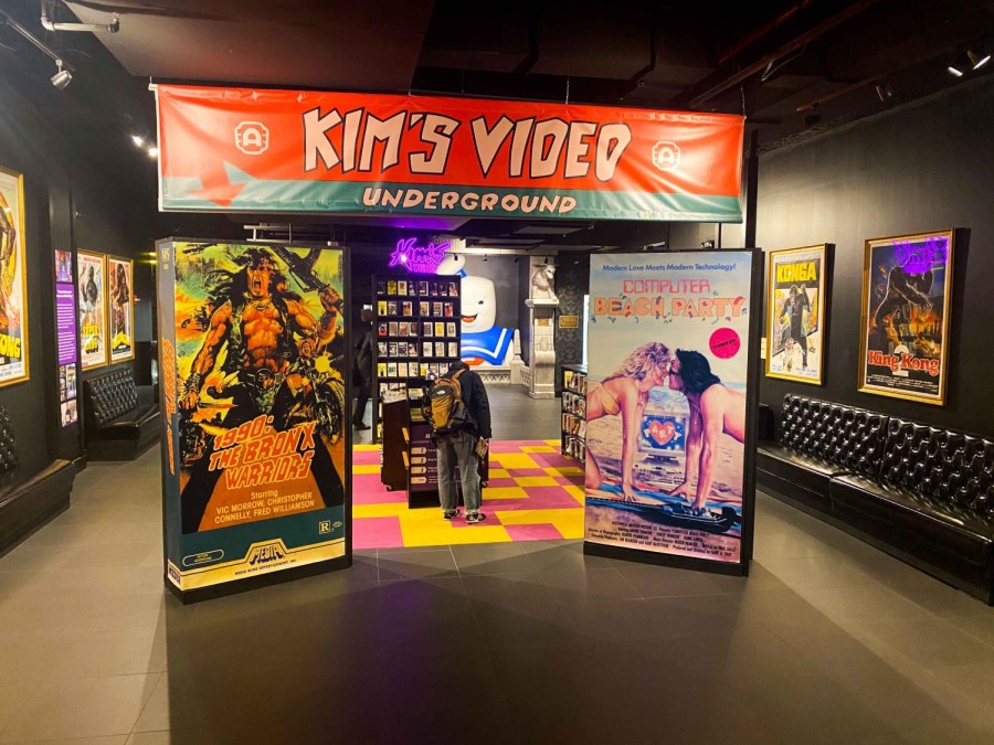 The indoor stand of Kim’s Video. The square structure stands in the middle of a hallway with framed movie posters. Inside the structure there are DVDs in display shelves.