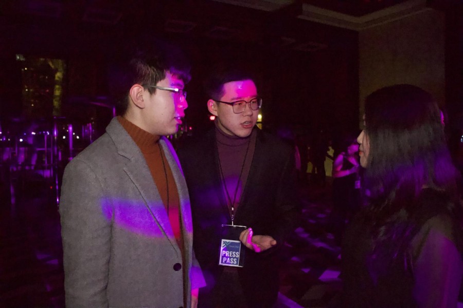 Three+gala+attendees+in+blazers+and+turtlenecks+chatting+under+the+purple+lights+of+the+V100+Gala+event.