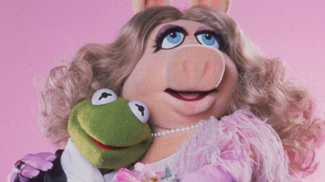 Miss Piggy is a stuffed puppet of a pink pig. A pink Muppet wearing a pink dress holds a green Muppet wearing a black-and-white tuxedo in front of a pink wall.