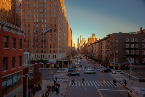 An elevated view of 10th Avenue as taken from the High Line at sunset. At the intersection in the foreground, cars and people zoom by.