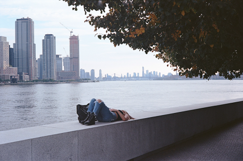 A woman lies on the barriers of the FDR Four Freedoms Park Conservancy on Roosevelt Island. On the right, a tree branches into the frame while in the back, the city skyline can be seen.
