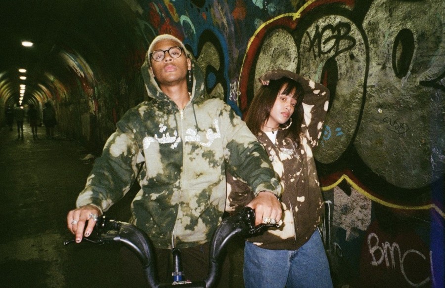 Two people stand in a tunnel with walls lined with graffiti. The one on the left is wearing a hoodie with green splotches on it, and is holding the handlebars of a bicycle. The one on the right is wearing blue jeans and a similarly colored splotch hoodie. People can be seen walking through the tunnel in the background.