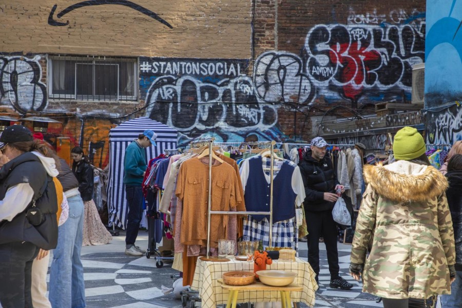 An anterior view of a lot the size of a New York City house filled with racks of clothes and tables for jewelry and other smaller items with graffiti on the wall in the background.