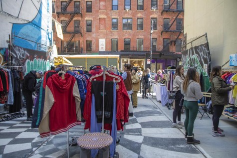 A posterior view of a lot the size of a New York City house filled with racks of clothes and tables for jewelry and other smaller items with a red brick building in the background.