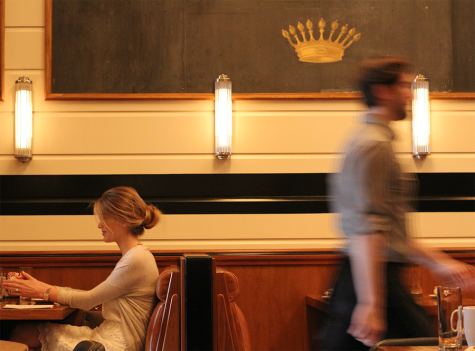On the left, a blonde woman sits at a restaurant booth. On the right, a blurred-out waiter dashes past. Above them, a blackboard with a gold crown hangs on the wall.
