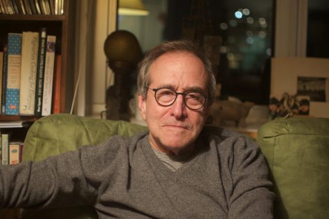 A headshot of NYU professor Mark Crispin Miller wearing a gray sweater. He is sitting on a green couch, next to a bookcase and in front of a window, during the nighttime.