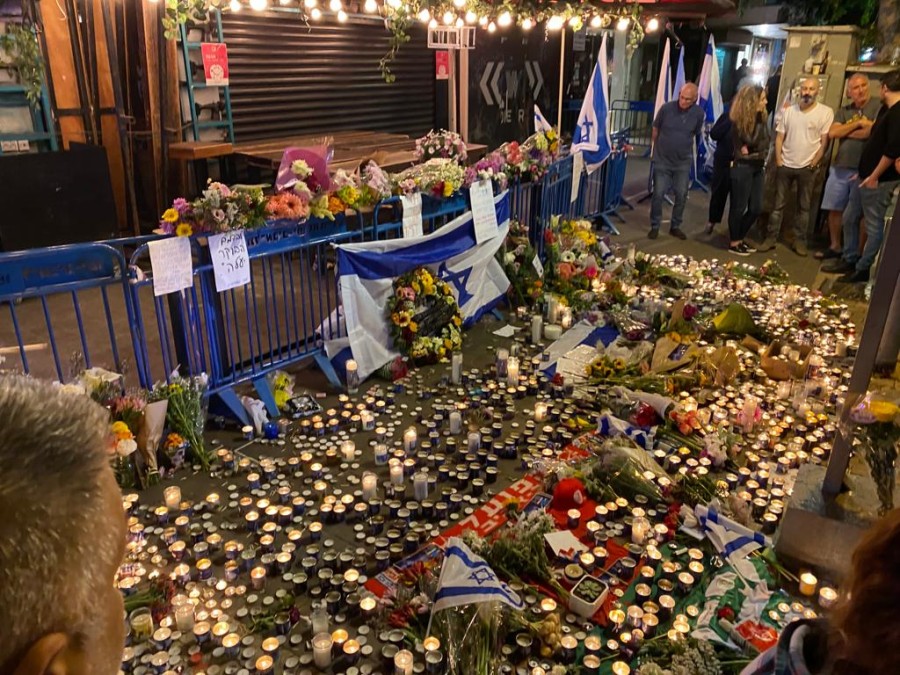 A crowd surrounds a memorial filled with candles, flowers, posters and Israeli flags in front of a storefront.