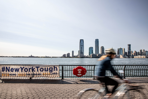 A blurred biker rides along the Hudson River Park piers. On the left, a white banner hangs on a rail with the words “#NewYorkTough” in royal blue. Behind both is the Jersey City skyline.