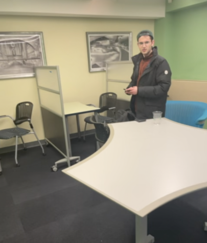 Unknown man in his 20s. He wears a green cap, orange sweater and a black down jacket. He is seen standing in front of a white desk in the Rubin Hall lounge while holding his phone.