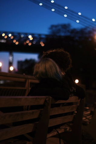 The backs of two people sitting together on a park bench in the early evening. Both look toward the East River.