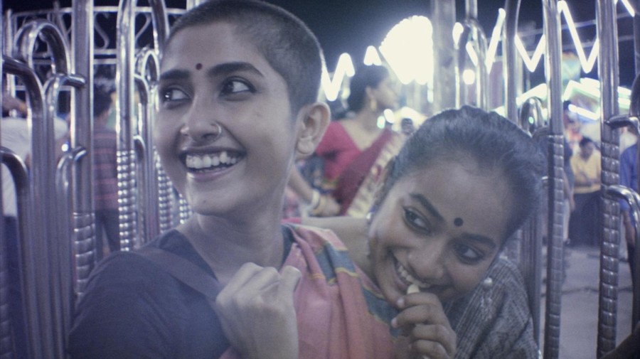 A still from the film of two women looking to the side and laughing. They are surrounded by metal bars on the side and are wearing saris.