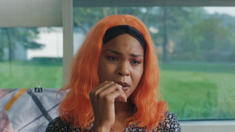 A woman with orange hair and a black headband wears a pensive expression. She has one hand next to her mouth and appears to be biting a bit of her nail. Behind her there is a window that looks out to green trees and grass.