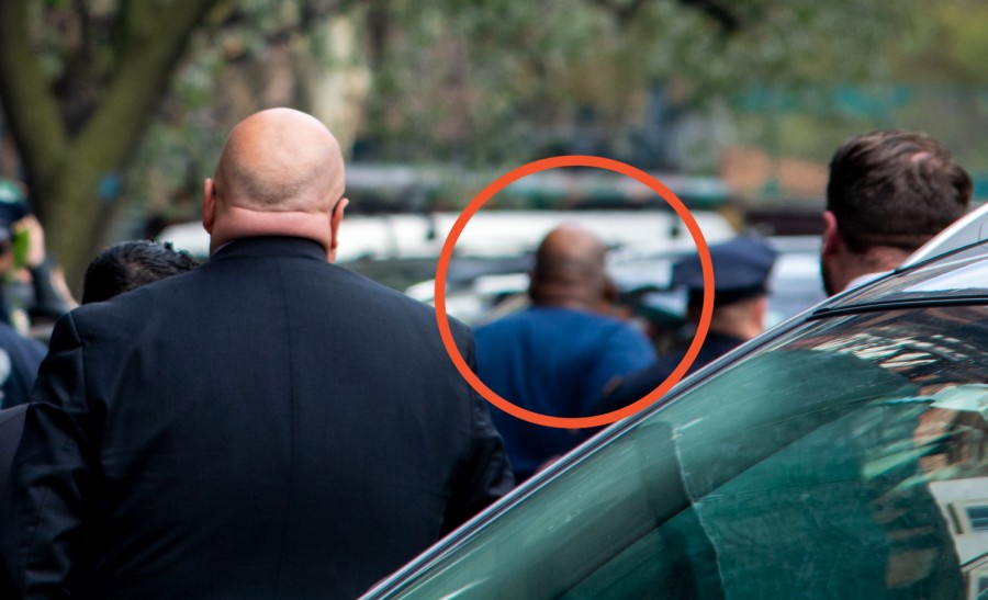 A close-up shot looking past the windshield of a car. A red circle inserted into the image highlights the head of Frank James, who can be seen as a blur in the background.