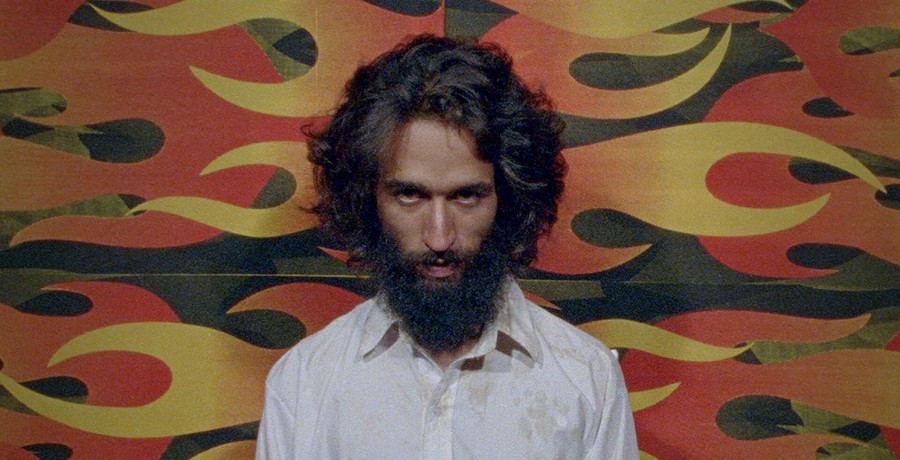 A man with a long beard and hair stares straight ahead, his eyes downcast. He’s wearing a plain white shirt and is against a background of painted sideways flames.