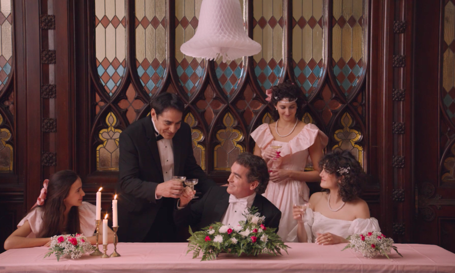 Three women wearing pink and white dresses gather around two men wearing black and white tuxedos. They toast their glasses in front of a stained-glass and wooden wall behind a table with a pink table cloth, flowers and lit candles.