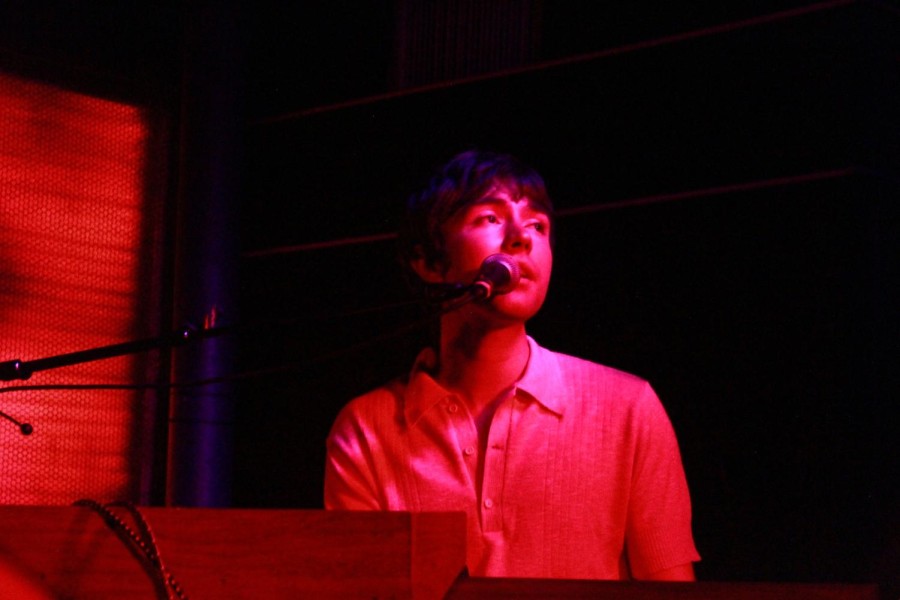 Matt Maltese sings into a microphone while sitting down and looking away. Red lights glow onto him and the stage.