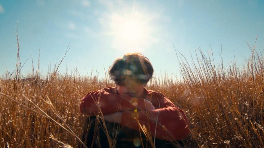 A person sits in a field of yellow wheat. Behind the person is a blue sky with the sun brightly beaming, creating flares that are cast down and blur the person’s face.