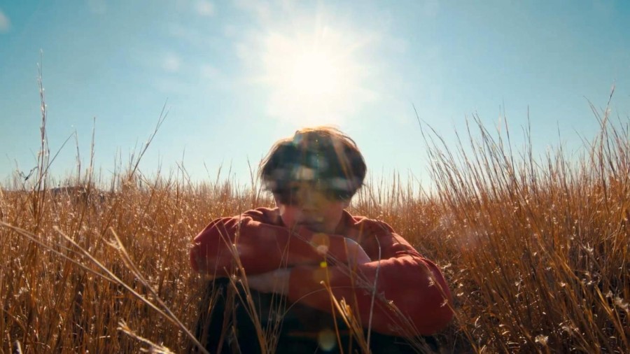 A+person+sits+in+a+field+of+yellow+wheat.+Behind+the+person+is+a+blue+sky+with+the+sun+brightly+beaming%2C+creating+flares+that+are+cast+down+and+blur+the+person%E2%80%99s+face.