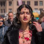 Portrait of Aarna Dixit wearing a peach-colored floral lehenga blouse with a black fur coat and traditional Indian gold jewelry. She is standing in Union Square Park’s farmers market looking confused, with a crowd of people behind her.