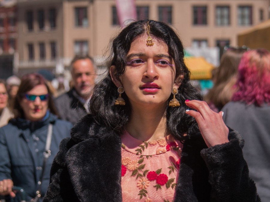 Portrait of Aarna Dixit wearing a peach-colored floral lehenga blouse with a black fur coat and traditional Indian gold jewelry. She is standing in Union Square Park’s farmers market looking confused, with a crowd of people behind her.