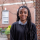 Portrait of Mika Chipana smiling and dressed in a white shirt with black polka dogs and a black sweater. She is standing in front of the red brick columns of the NYU School of Law courtyard, her hair neatly braided and flung to the back.