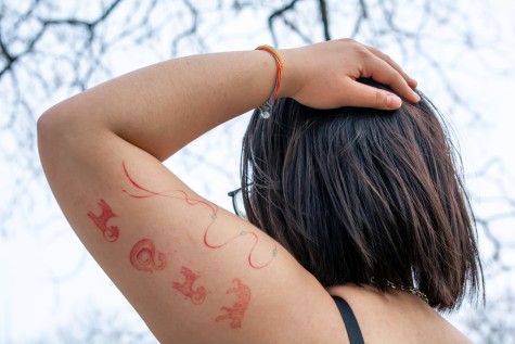 The back of Mayee Yeh’s left arm as they rest their hand on her head. The focal point of the photo is the various red tattoos on their upper arm.