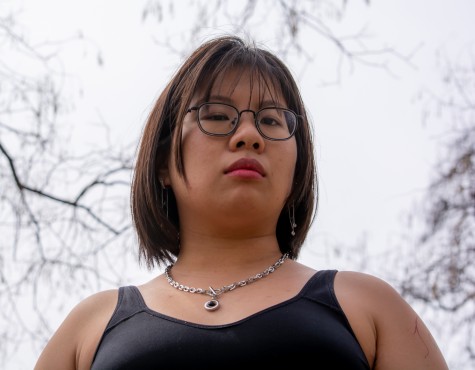 Portrait of Mayee Yeh. They are wearing glasses and a black tank top with a silver necklace. They are standing in a park during the daytime.