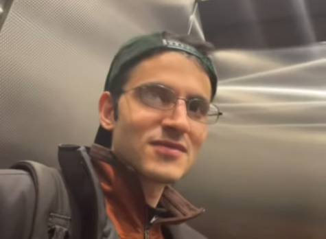 A close-up photo of a man in an elevator. He has light skin and glasses, and wears a green baseball cap backwards. He is wearing an orange jacket under a dark gray coat.