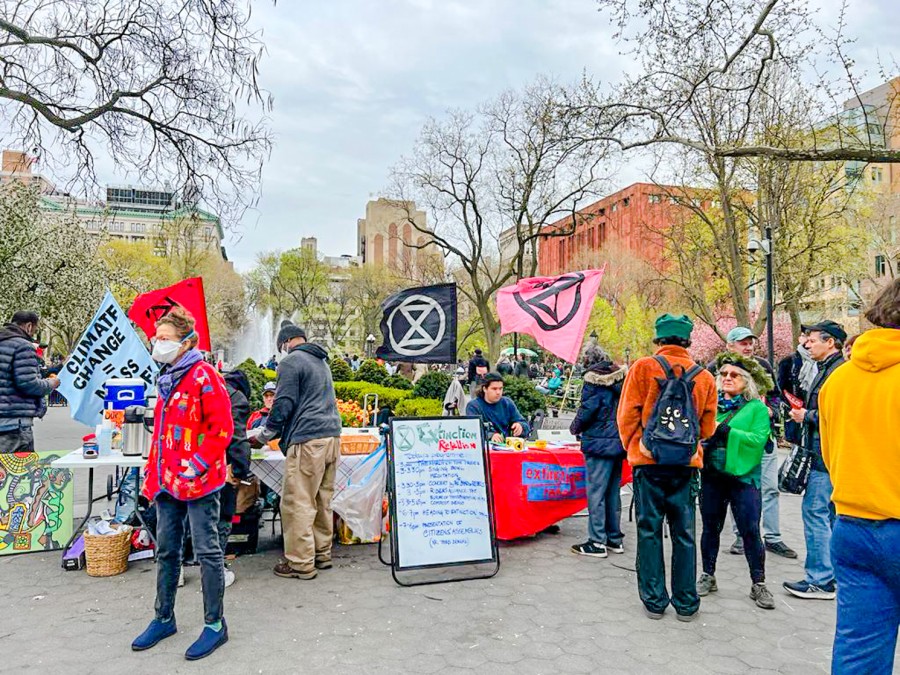 An+open+area+on+the+westside+of+Washington+Square+Park+surrounded+by+trees.+People+congregate+around+Extinction+Rebellion+NYC+tables+and+banners.