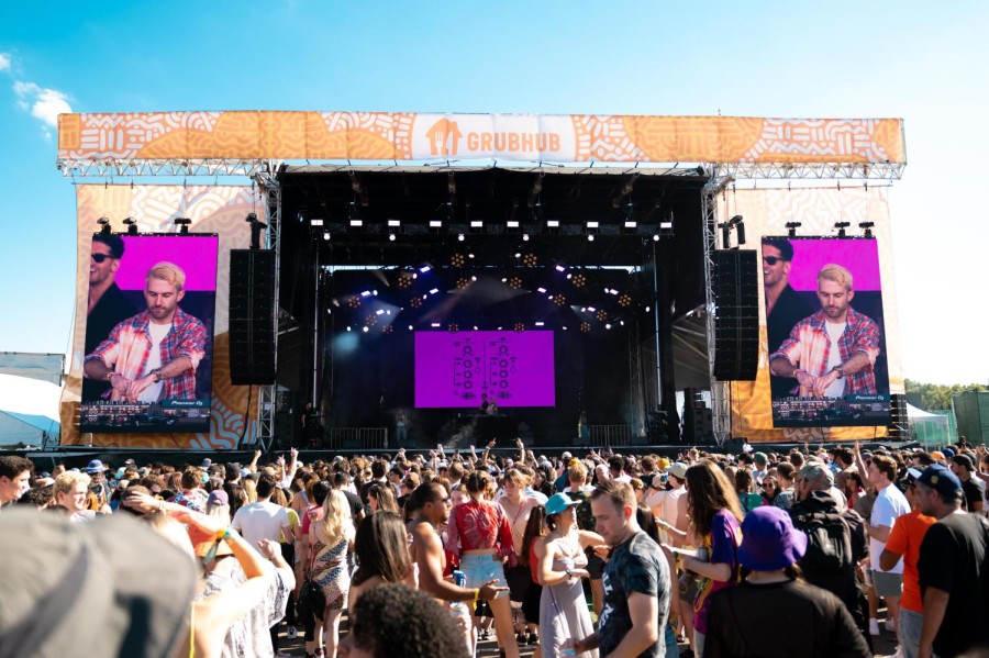 A crowd of concert-goers dressed in summer outfits in the foreground and an outdoor stage in the background. The weather is sunny.