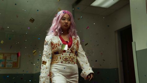 Michelle Yeoh walks through a hallway surrounded by falling pink, yellow and blue confetti. She has pink hair and wears a white bedazzled jumpsuit.