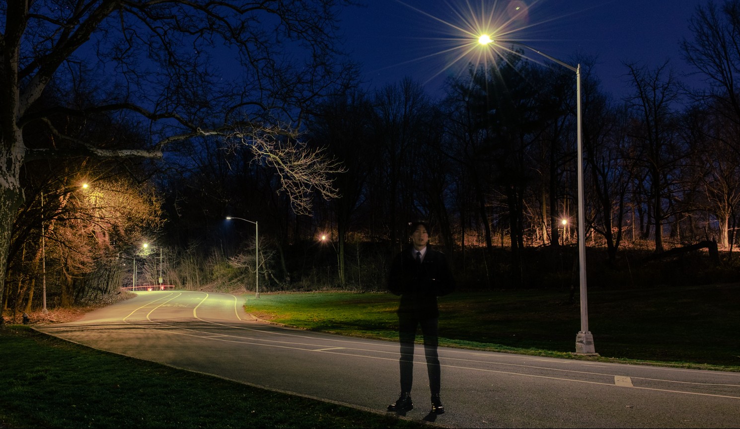 A male-presenting figure stands on a road at night surrounded by trees.