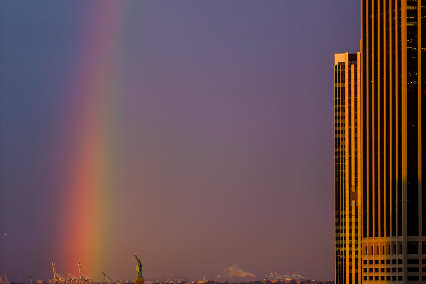A rainbow to the left; the Statue of Liberty at the bottom and white office building cast in golden sunrise light to the right.