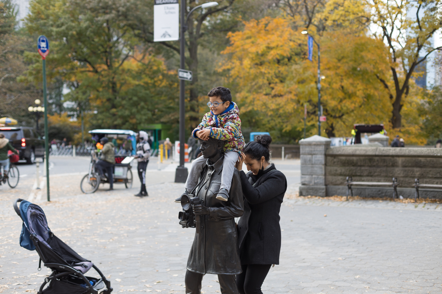 A life-size bronze statue of Diane Arbus with a male-presenting child sitting around Arbus’s shoulder. A female-presenting adult is holding the child with both hands.