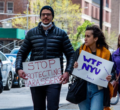 A man and a woman stand side by side as they walk on the sidewalk. They hold signs that read, from left to right, “Stop Protecting Harassers” and “WTF NYU.”