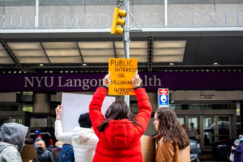 A girl in a red jacket holds a yellow poster that reads “Public Interest or Billionaire Interests.” In front of her is the building of NYU Langone Health.