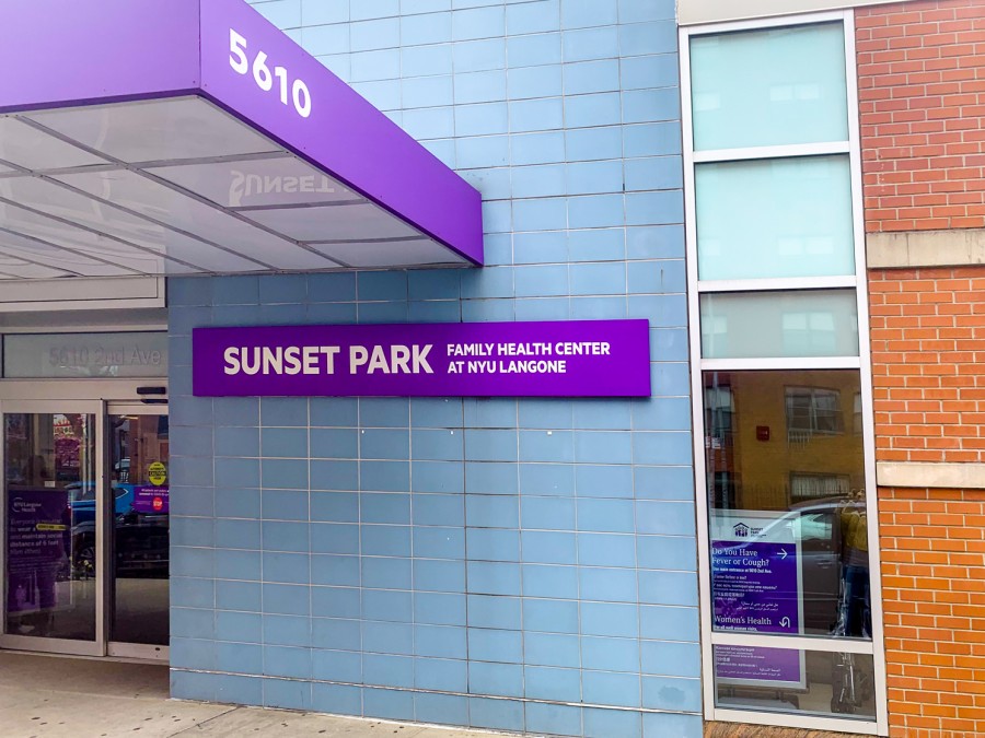 The text “Sunset Park Family Health Center at NYU Langone” is printed in white on a purple plaque against a blue wall on the front facade of the NYU Langone Sunset Park building.
