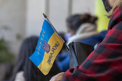 A close-up image of a blue-and-yellow Ukrainian flag. In the blue part, the word “Ukraine” is written in Ukrainian in yellow text. In the yellow part, the country’s name is written in English in blue text. On the right, a person’s arm extends into the frame. They are wearing a red-and-black flannel.