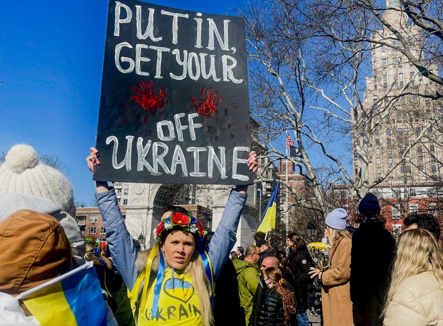 A person in Washington Square Park wears a yellow shirt that reads “Ukraine” and holds a sign above their head reading “Putin get your,” then two red handprints, “off Ukraine.” In the background, people hold Ukrainian flags at the park.