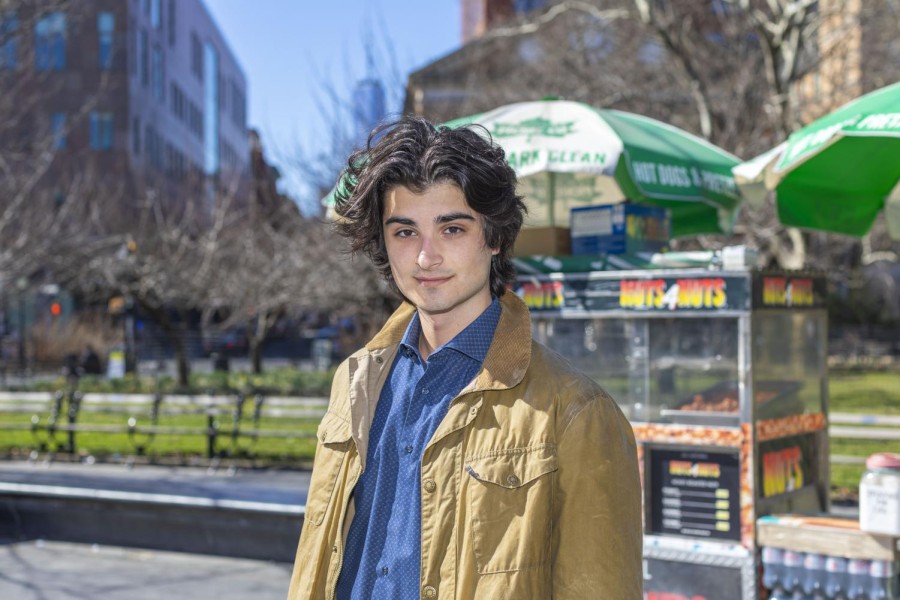A portrait of Edoardo Marras with long wavy hair wearing a blue shirt and beige coat in front of the halal cart at Washington Square Park.