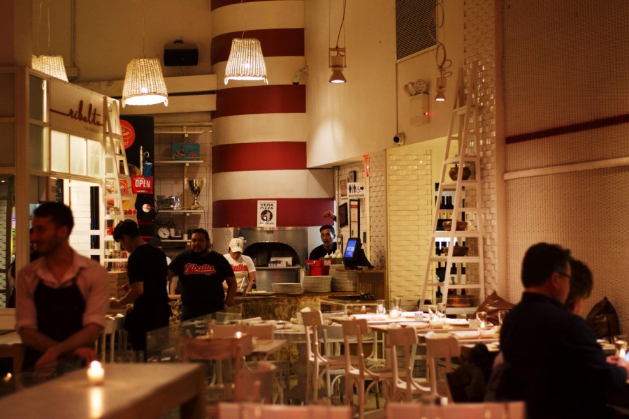 Interior of Ribalta with white walls to the side and red-and-white striped walls at the back. In the foreground are white chairs and tables, with two diners and one waiter visible. In the background is a pizza oven and a plaque with the Ribalta logo in red. The room’s lighting is warm and dim.