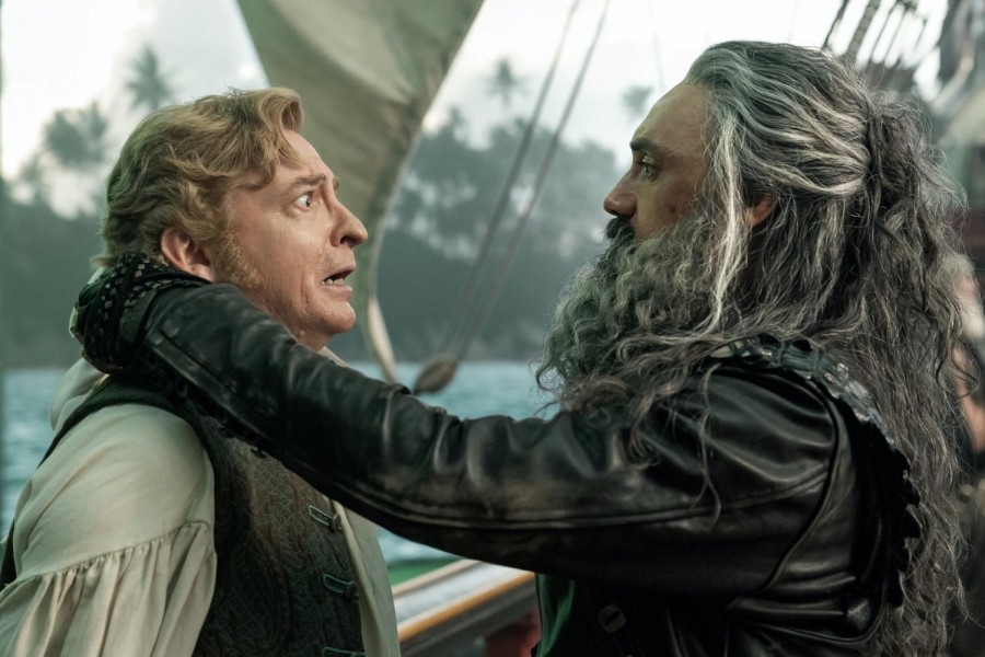 Actor Taika Waititi has black and gray hair, a long beard and a black jacket. His left hand is holding actor Rhys Darby by the neck. Darby has blonde hair and has his mouth opened in a panicked expression.