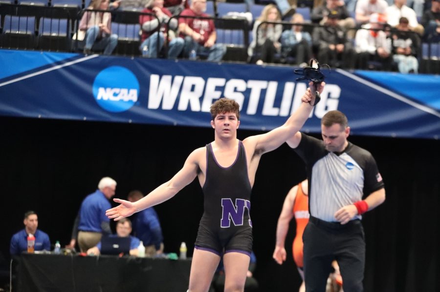 Cooper Pontelandolfo and an NCAA referee stand in the center of the frame. An NCAA Wrestling banner runs through the length of the background, below spectators in the bleachers.