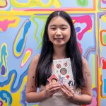 Portrait of Grace Xiang holding up an assortment of stickers. Xiang is standing in front of an abstract multicolored background.