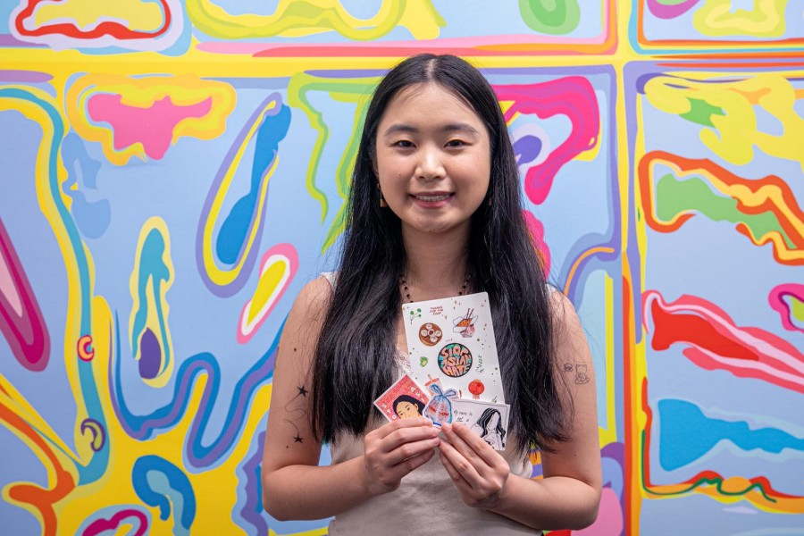 Portrait of Grace Xiang holding up an assortment of stickers. Xiang is standing in front of an abstract multicolored background.
