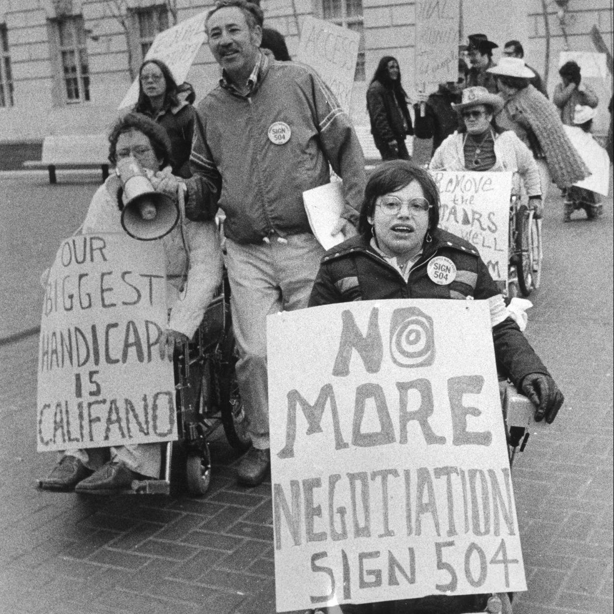 A black-and-white picture of Judith Heumann at a protest holding a sign that reads, "No more negotiations, sign 504." Other people attending the protest are behind Heumann in the background. One protester holds a sign that says "Our biggest handicap is Califano," while a man holds a megaphone up for them to speak.